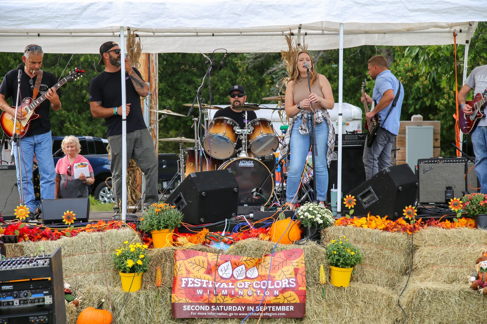 Band with female lead singer performs on stage under white tent with hay bales, pumpkins, and mums decorating a stage.