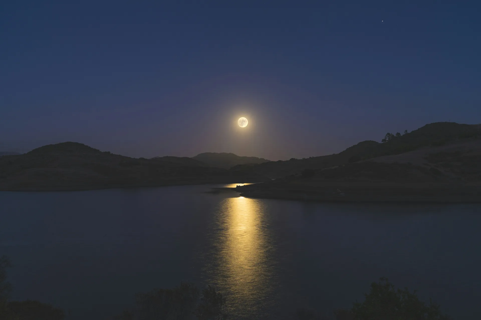 Full moon in dark sky over lake and trees shoreline hills with moon reflecting in the water