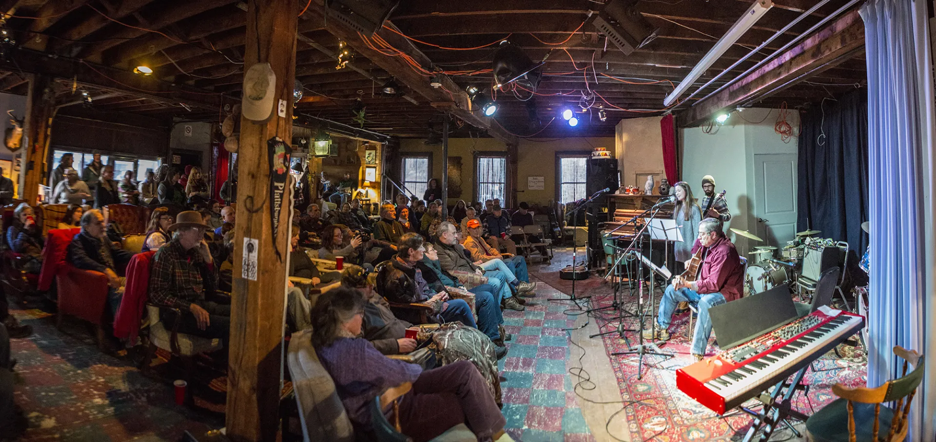 Man plays guitar on stage at Upper Jay Art Center with full house of people sitting on various chairs and couches