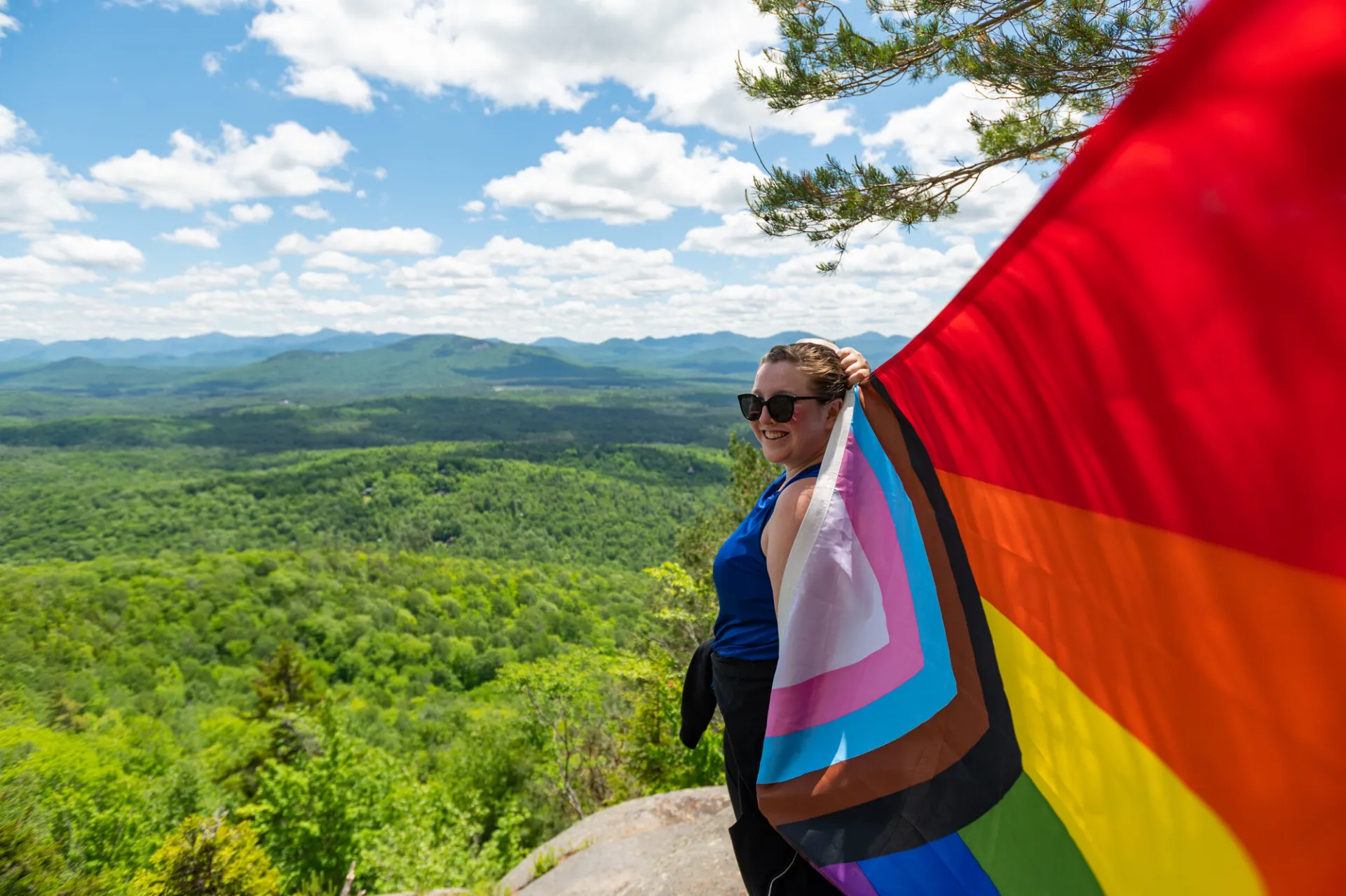 Person in hiking clothes and sunglasses stands on top of a mountain in summer holding pride flag billowing behind them