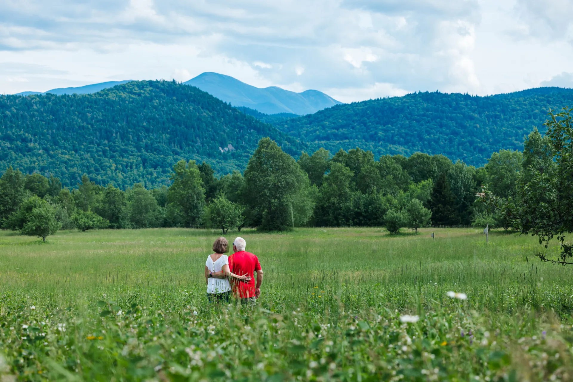 Senior couple stands in field of tall grass with arms around each other's waists looking at mountain view