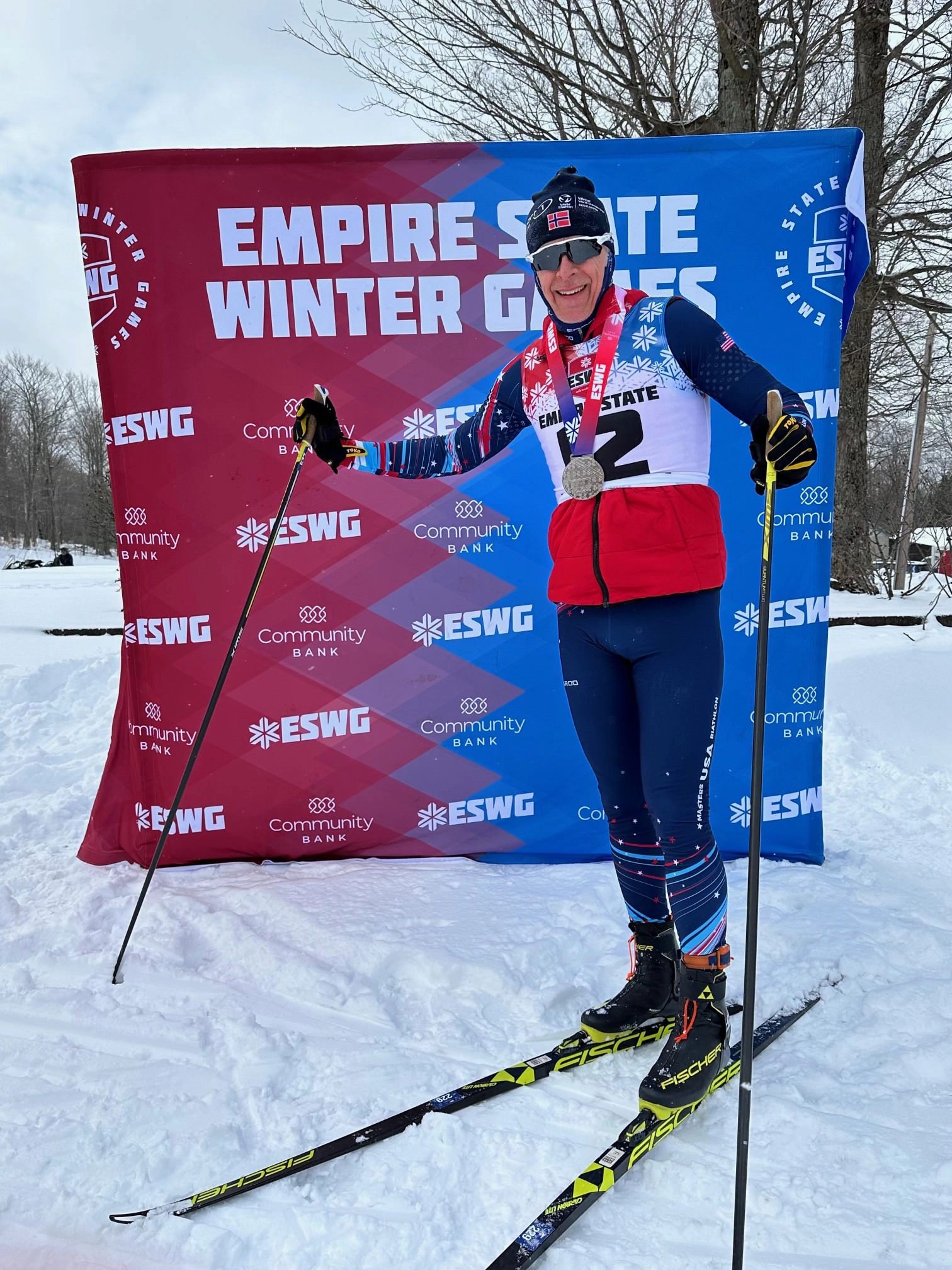 Skier with medals and ski poles poses proudly in front of a branded Empire State Winter Games backdrop.