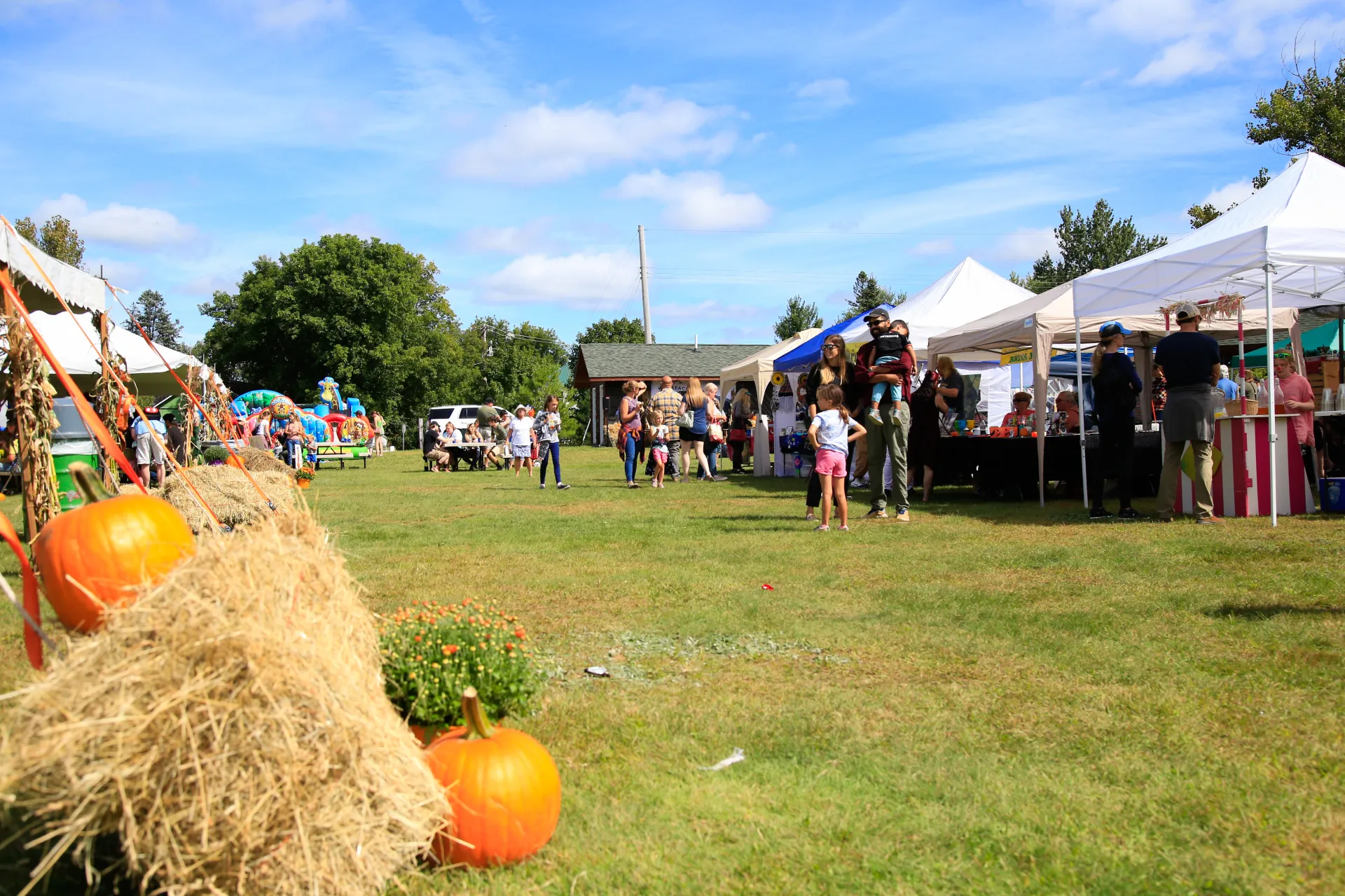 People browse at vendor tents in the background at outdoor event with pumpkin and hay bales in foreground