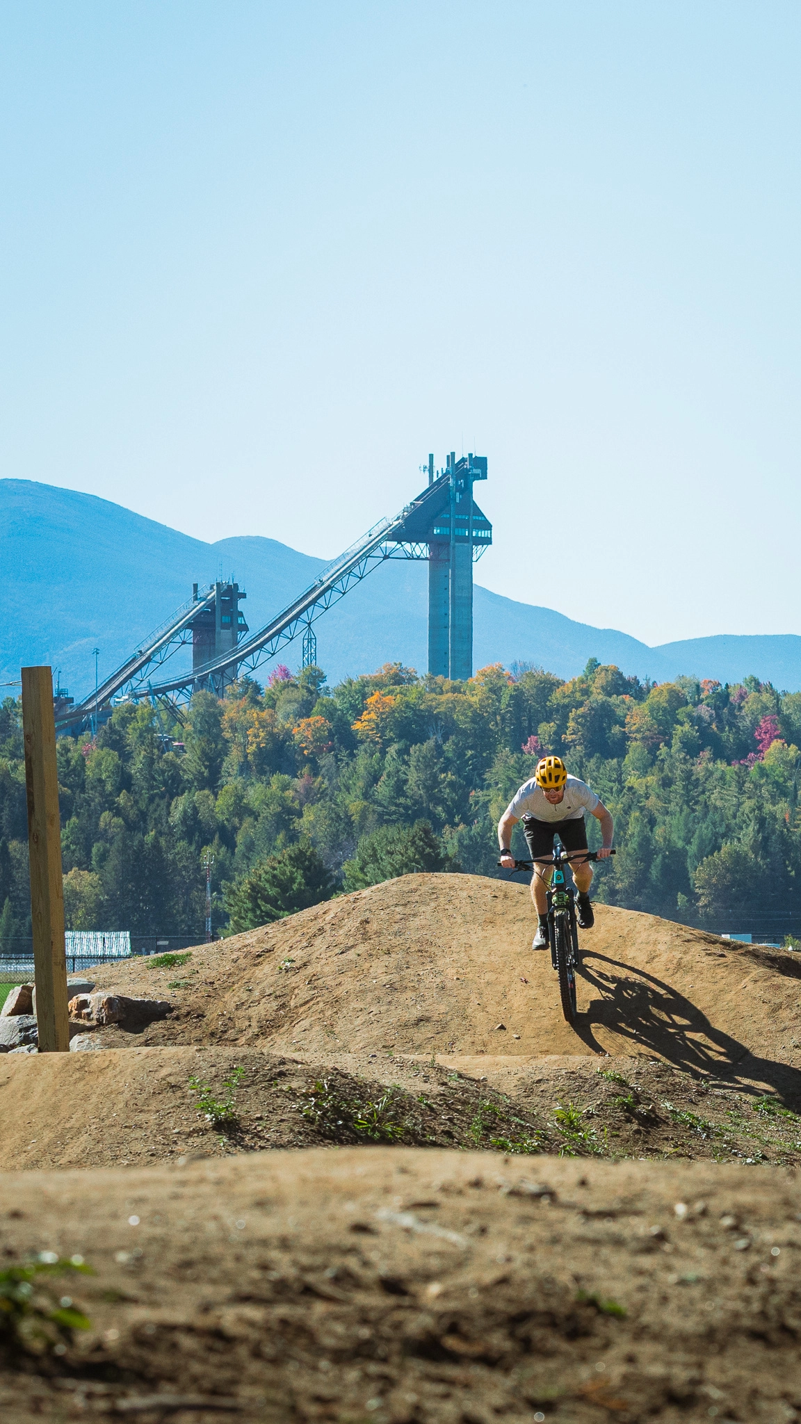 A man rides a mountain bike over a dirt hill with mountains in the background.