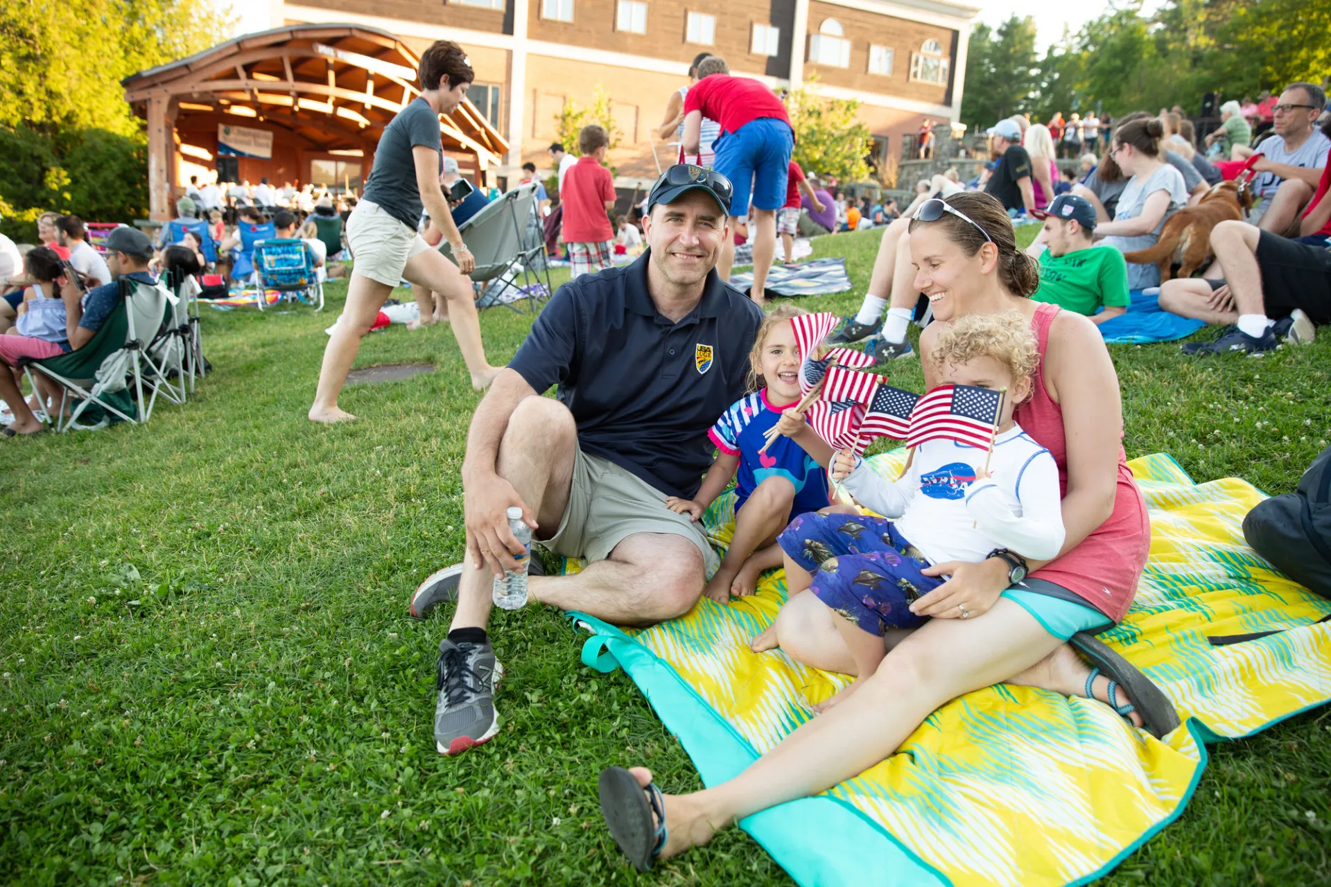 A family sits on a large towel in the grass and waves American flags.