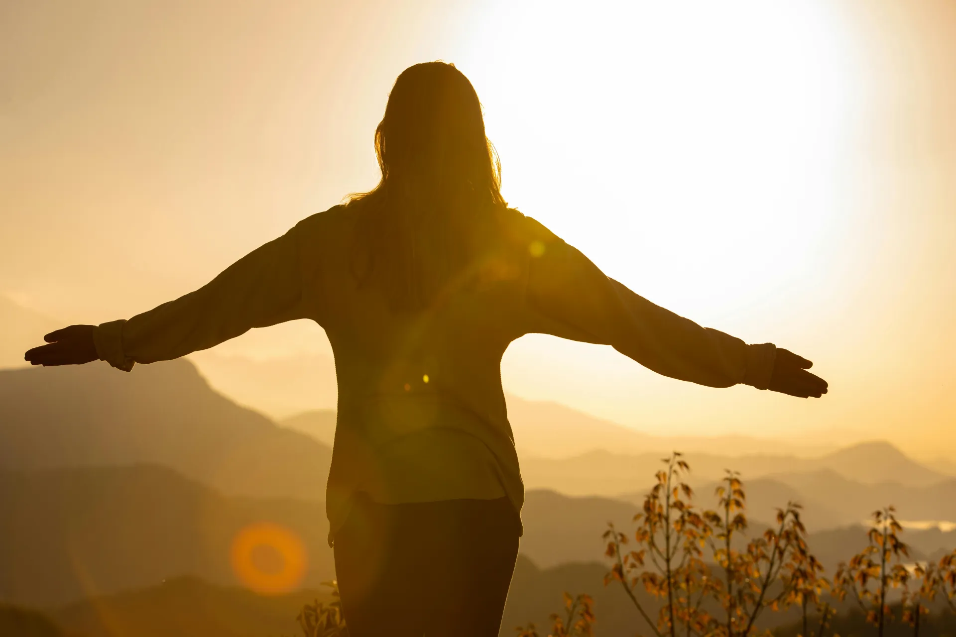 Woman's silhouette with outstretched arms in front of distant mountain view at golden sunrise.