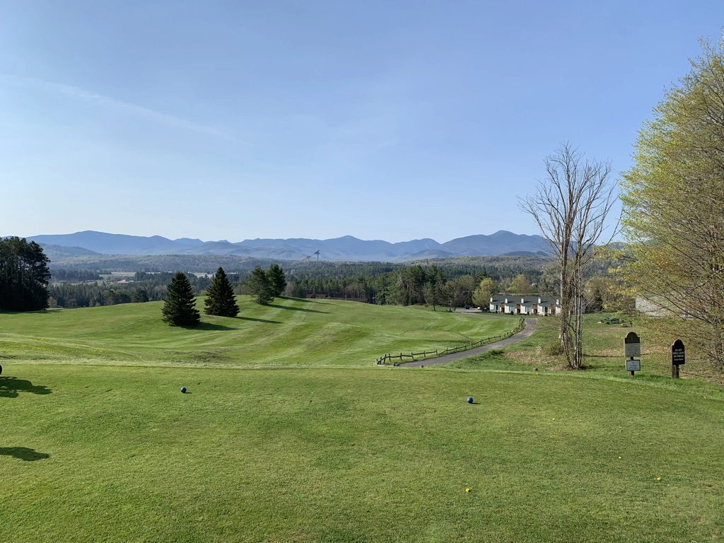 Tee and fairway at Lake Placid Club golf course, with Olympic ski jumps and Adirondack peaks in the distance.
