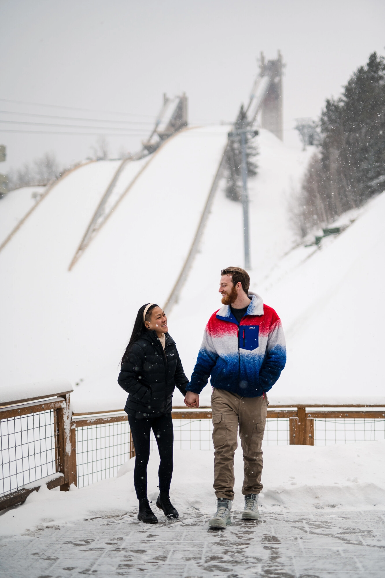 A couple holds hands and smiles at each other in front of snow-covered ski jumps.