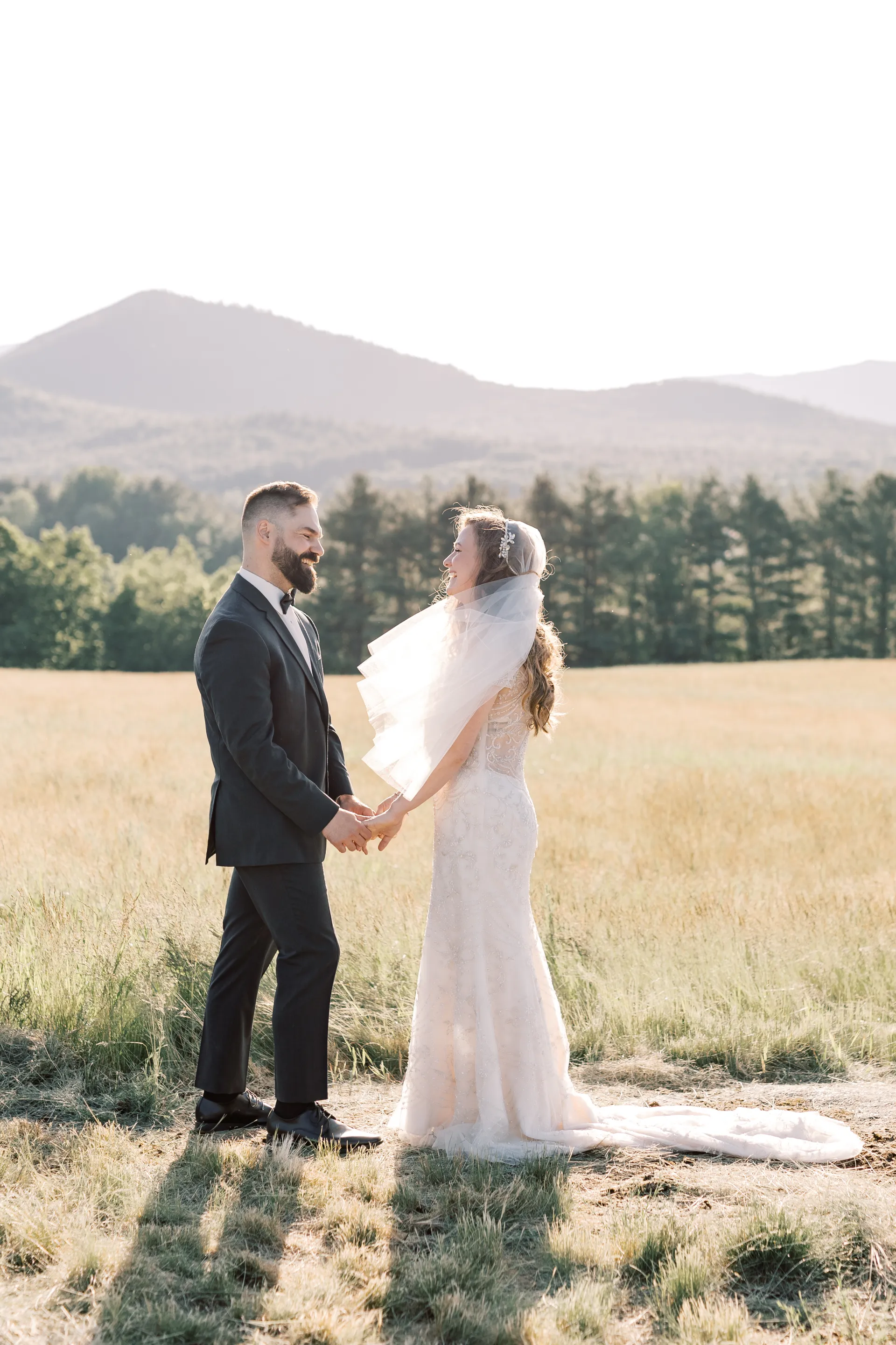Bride and groom smile at each other in a wheaty field with veil blowing in the breeze