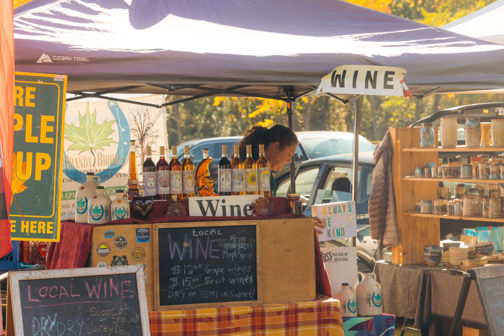 Woman holding signs that says "always be kind" stands behind wine display decorated with chalkboard signs under purple tent at farmers market