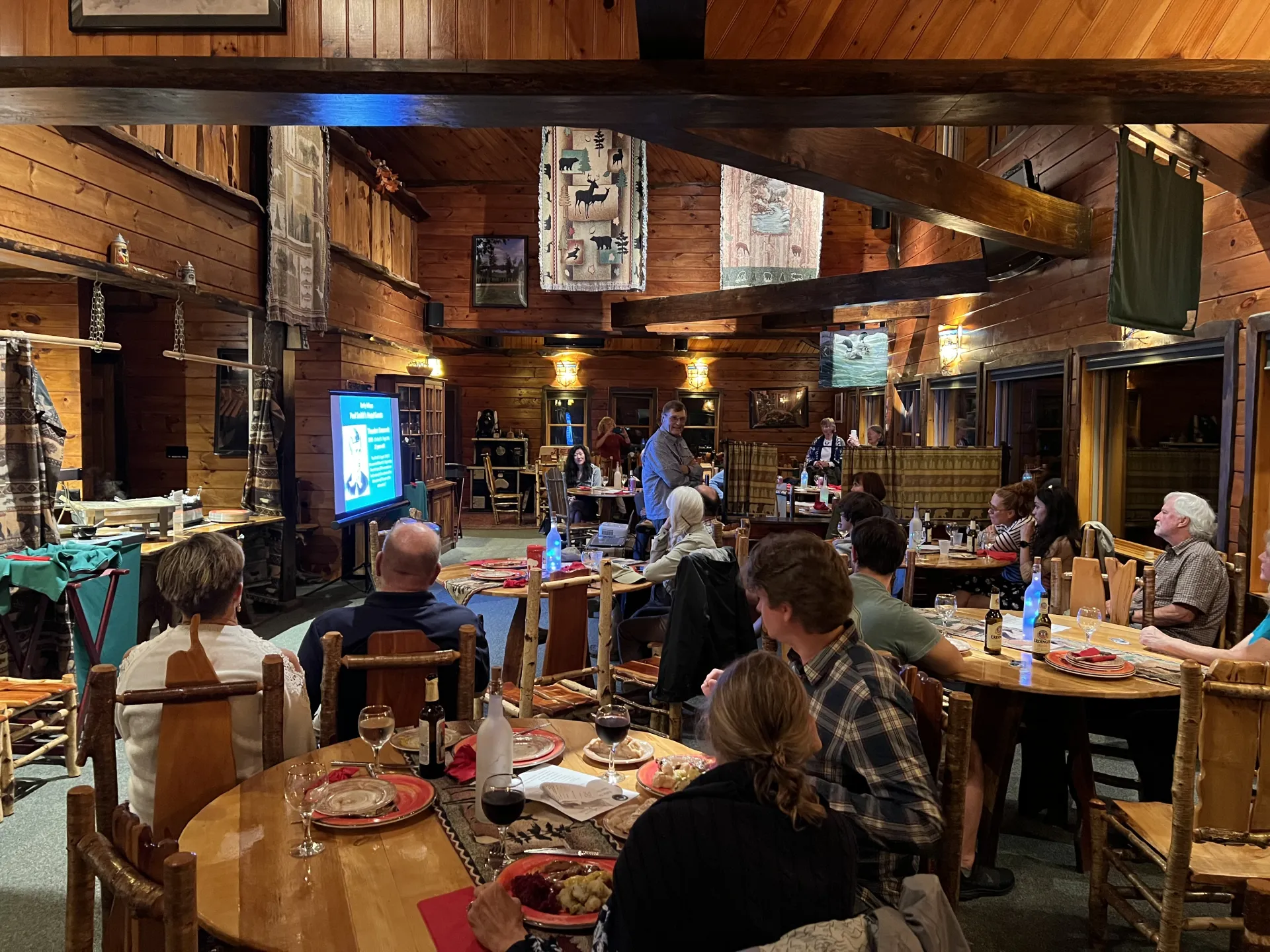 A group of people seated at round tables in a rustic wooden lodge listen to a presentation shown on a screen.