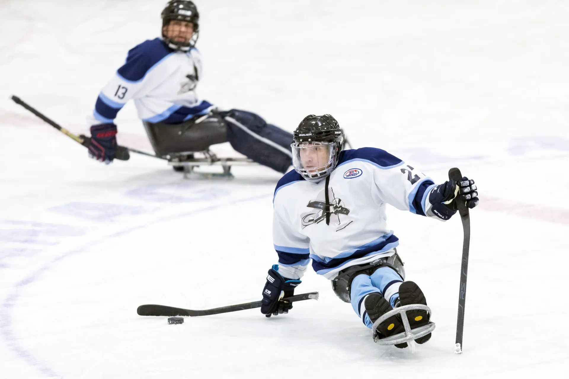 A close-up action shot of two para ice hockey (sled hockey) players on an indoor rink during the Empire State Winter Games.