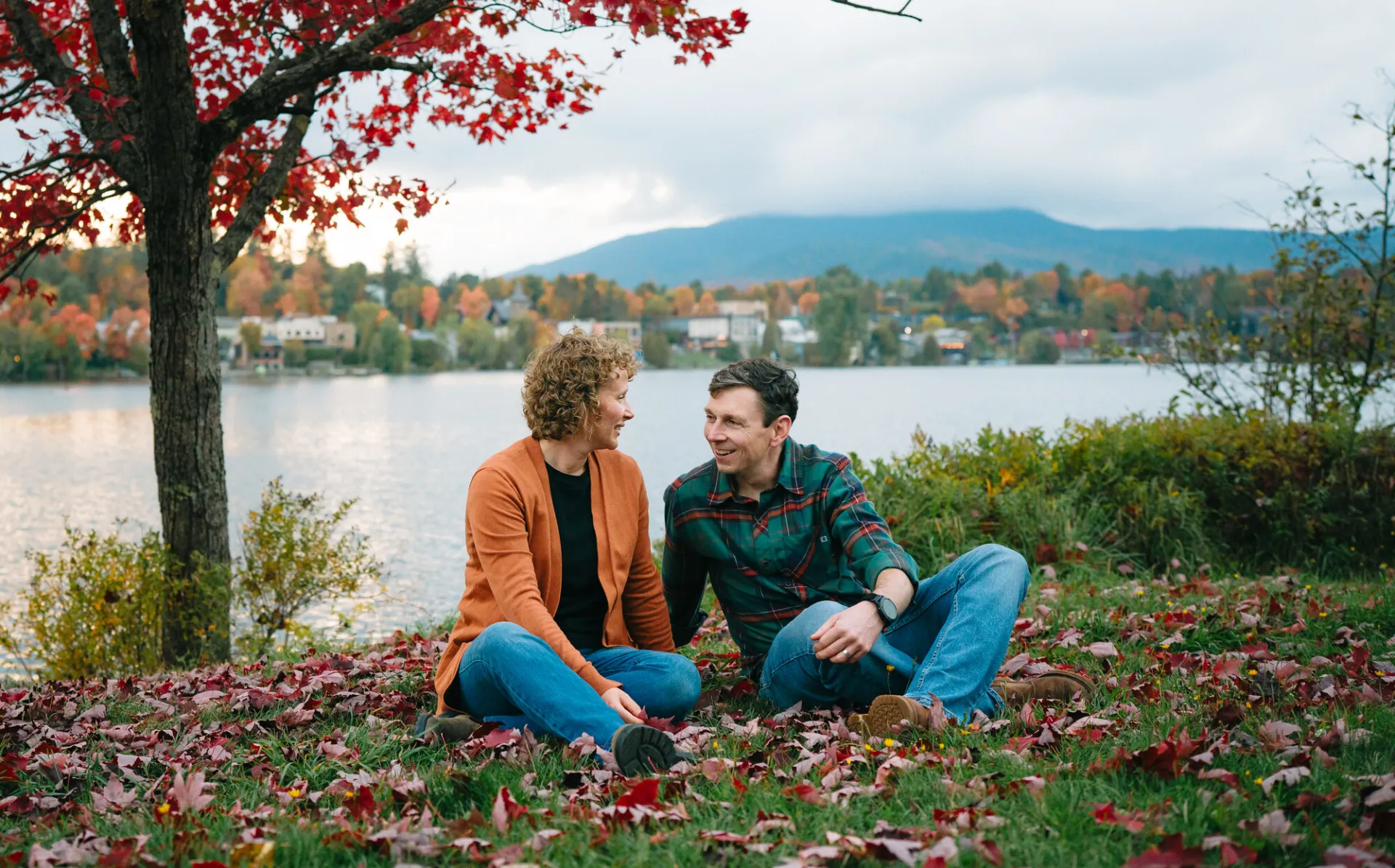 Middle-aged hetero couple sits on grass beside Mirror Lake on fallen leaves beneath tree with red leaves