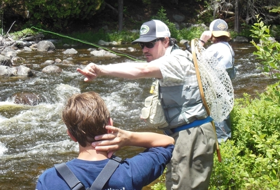 A man wearing a fishing vest and a baseball cap points across a rocky river while instructing a person in waders.