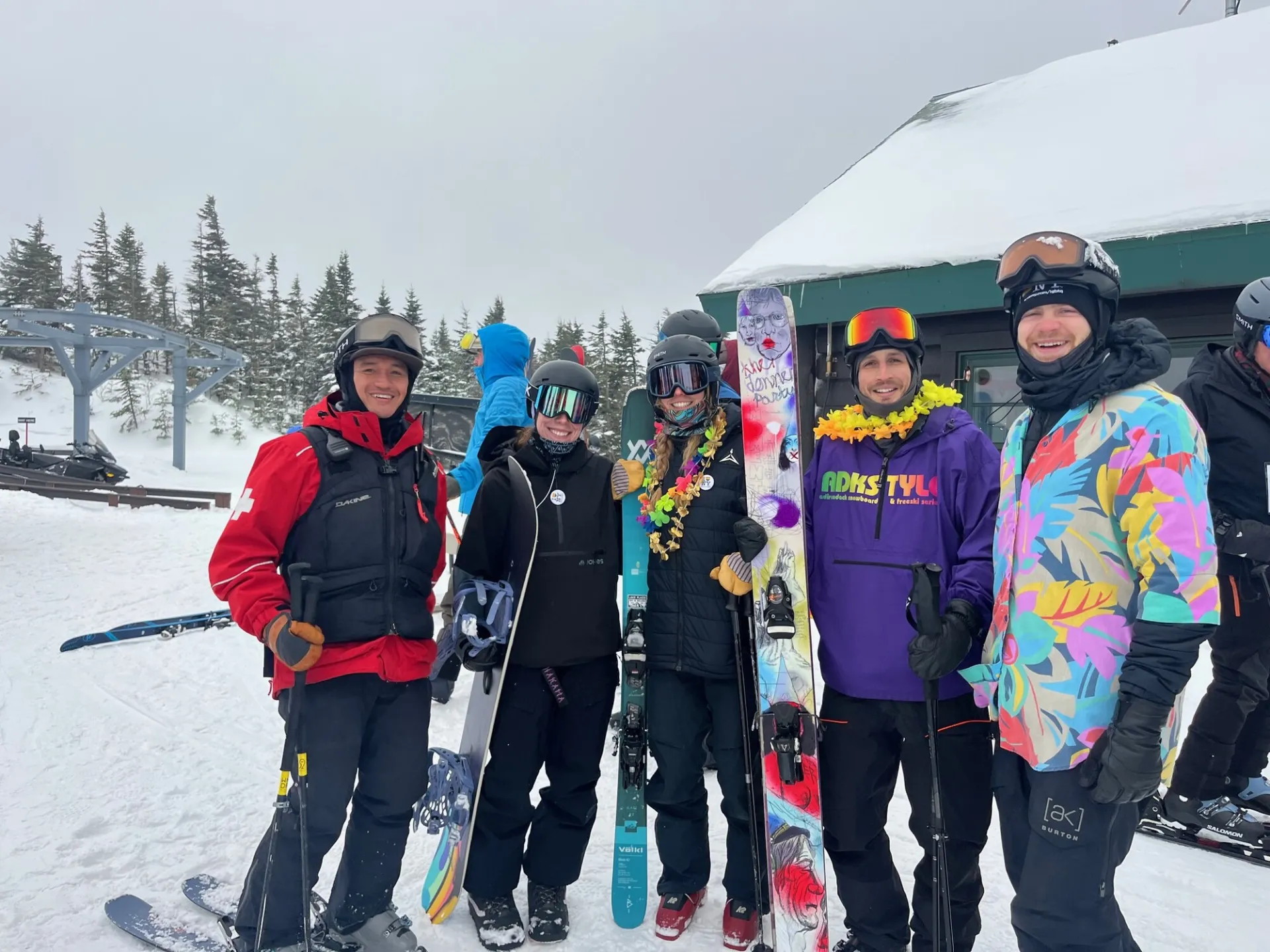 A group of skiers and snowboarders, some wearing colorful leis, pose together near a mountain building.