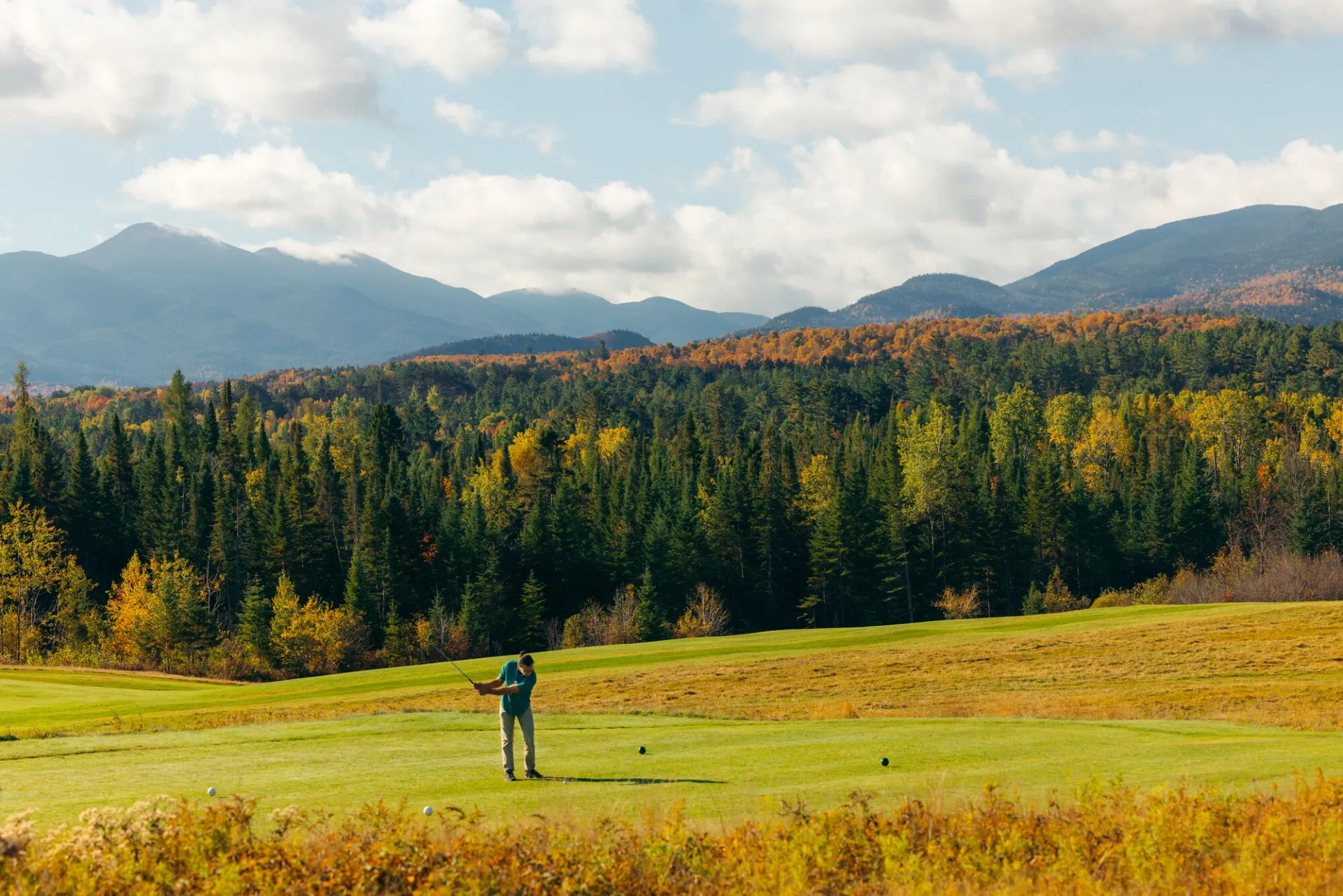 A golfer tees off on a green course, with soaring fall foliage-covered mountains in the background.