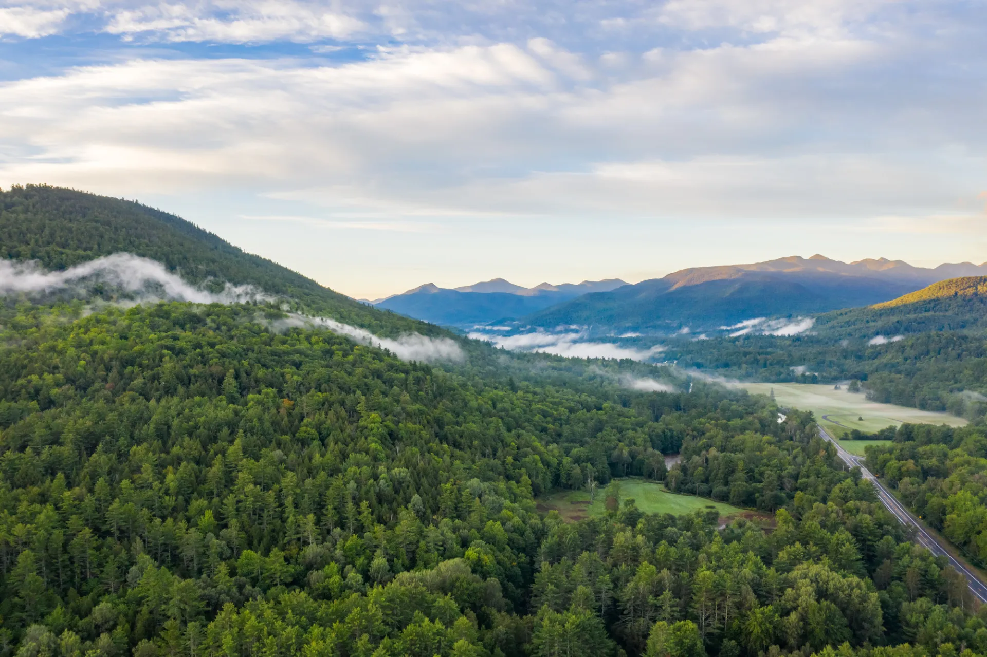 An aerial view of a forested mountain with a road and more mountains in the distance. 