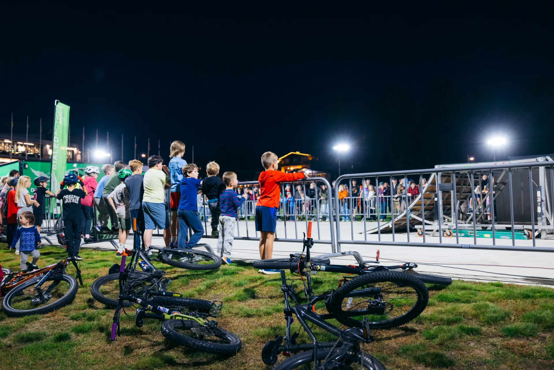 Bikes litter the lawn and children stand against event fencing watching BMX stunt show on Olympic Oval