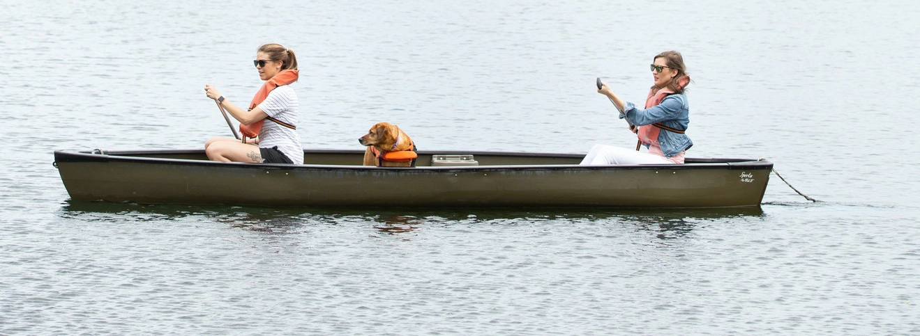 Two women and a dog in a brown canoe.