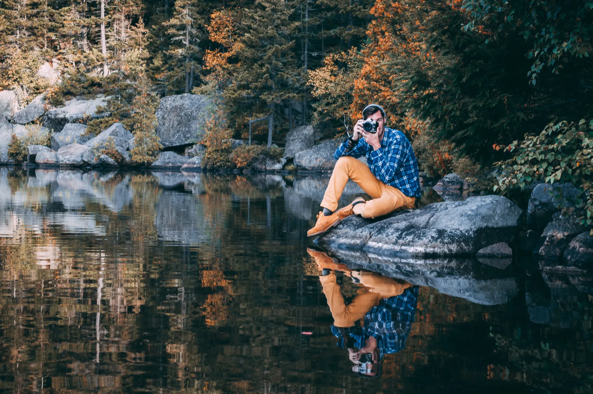 A person siting on a rock by the water holding a camera with fall foliage behind them.