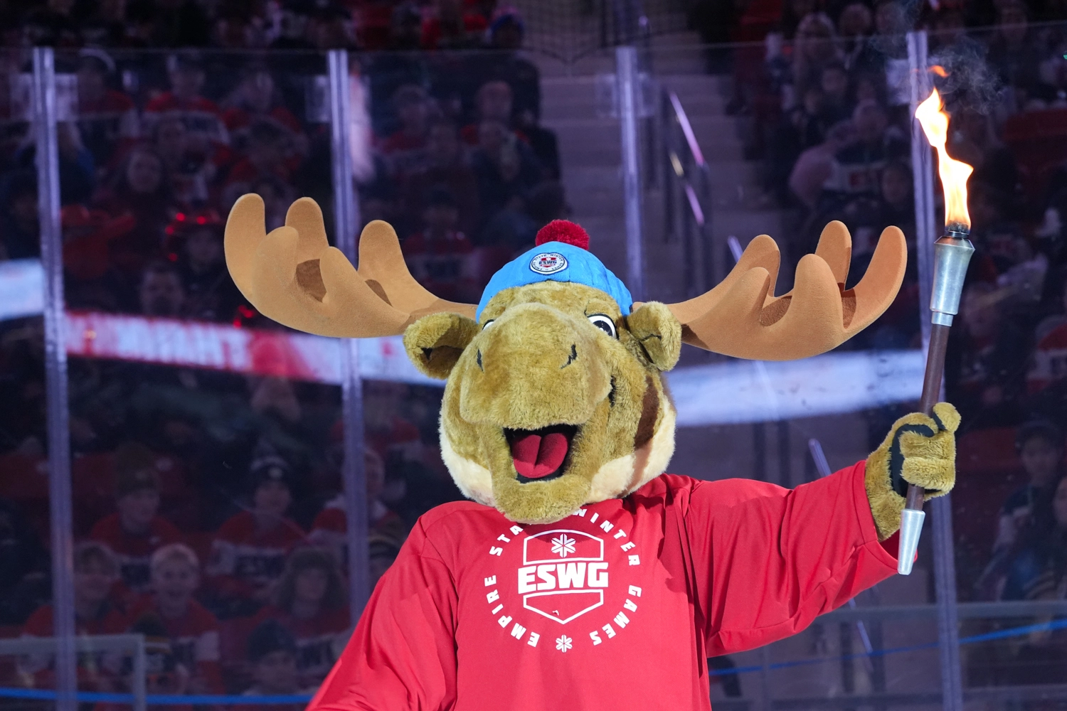 A moose mascot wearing a red Empire State Winter Games jersey holds a flaming torch aloft during an opening ceremony.