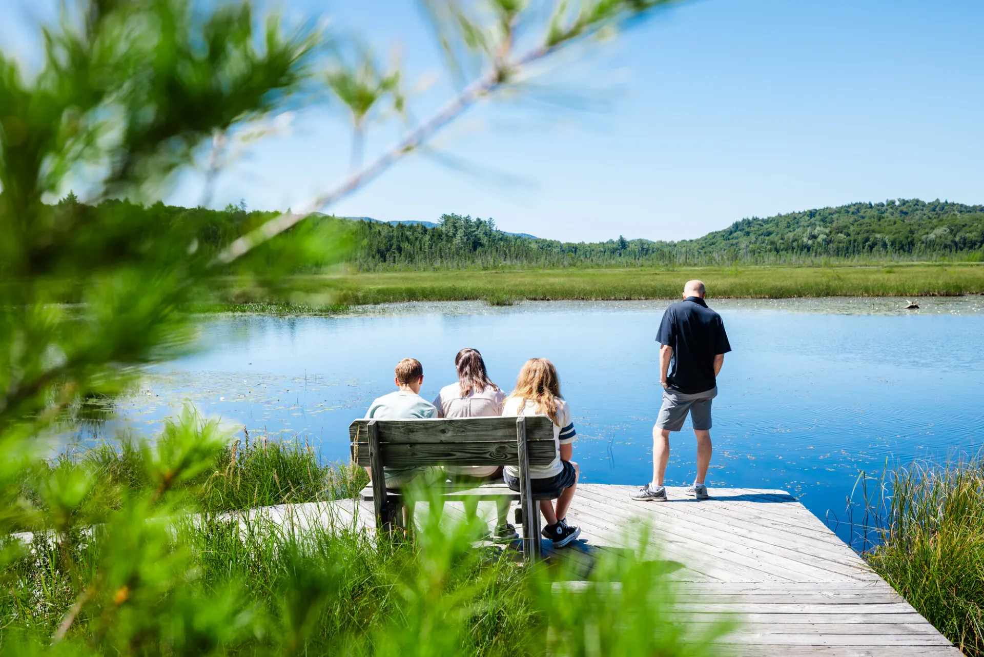Three people sit on a bench overlooking a pond while another person stands on a deck next to them.