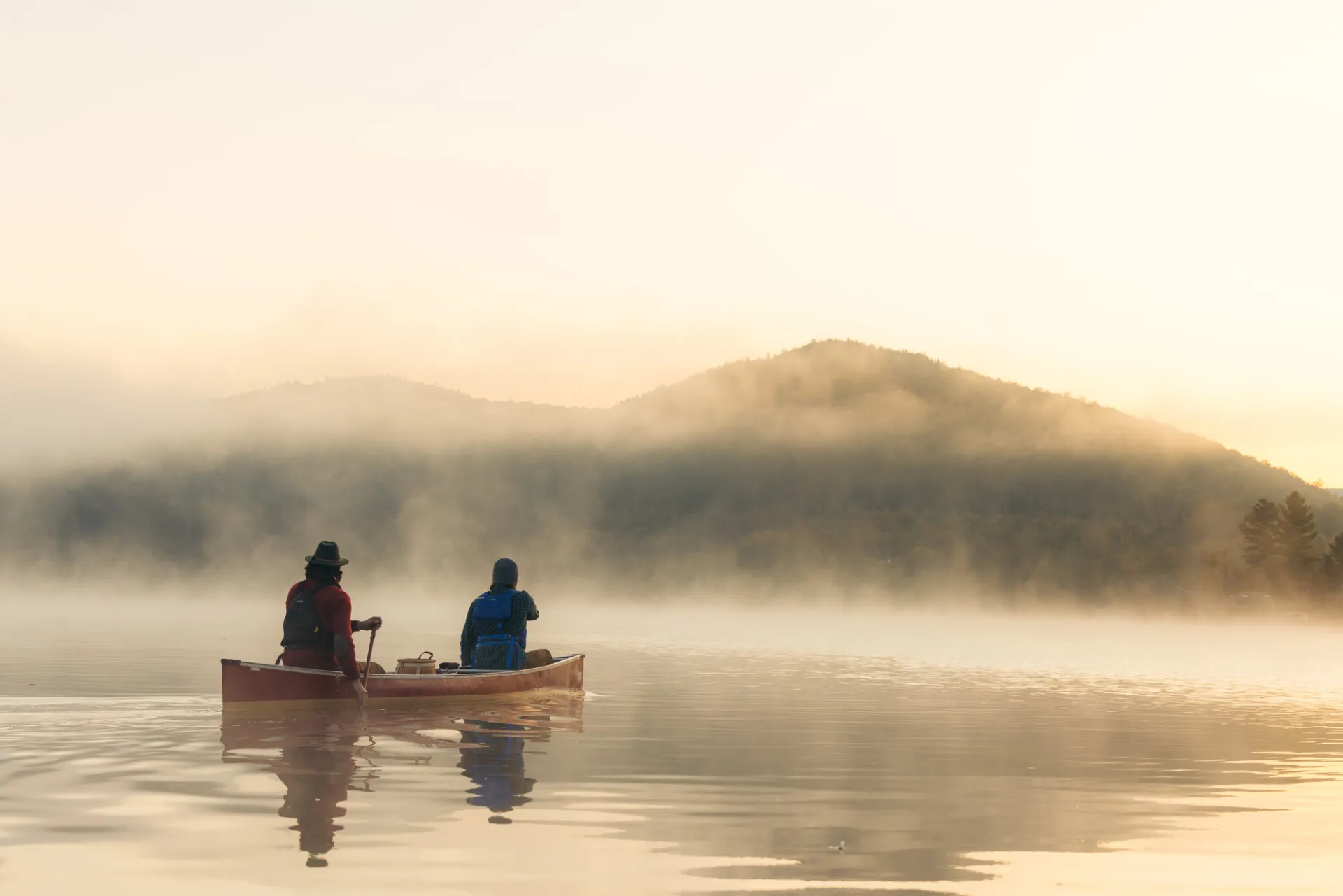 Two people, one wearing a wide brimmed hat, paddle away from the camera on a lake toward mountainous shoreline on a misty morning
