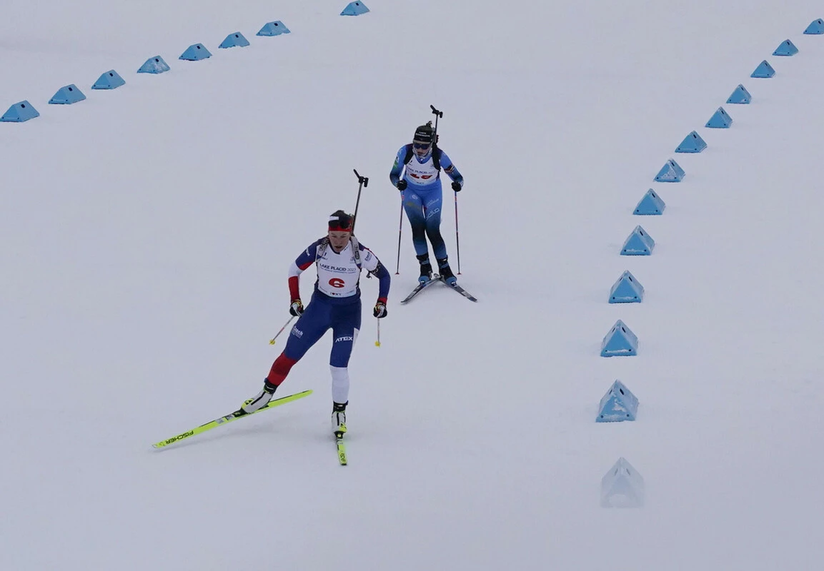 The image shows two athletes skiing on a snowy course marked by blue triangular markers.