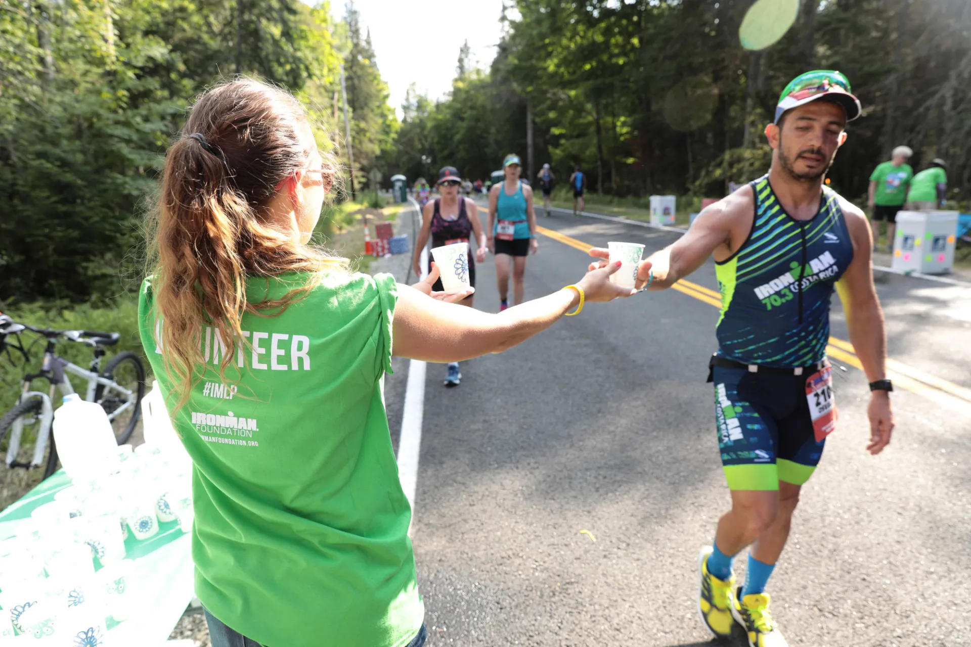 A woman with a ponytail wearing a green Volunteer tshirt is seen from behind handing a cup to an Ironman athlete while other athletes approach on the road behind