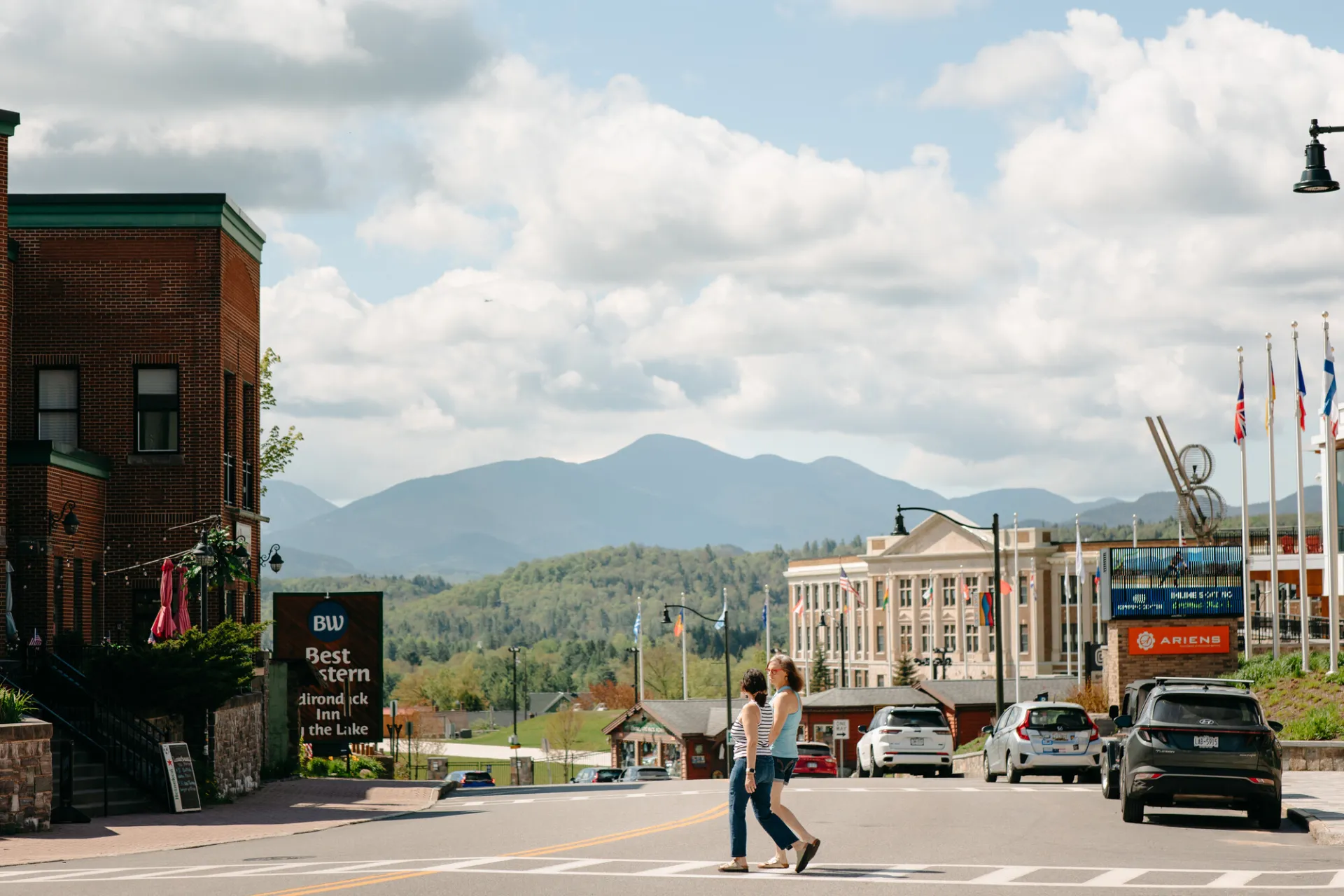 Two women walk across road in sidewalk with downtown businesses on either side and mountain views in the distant background