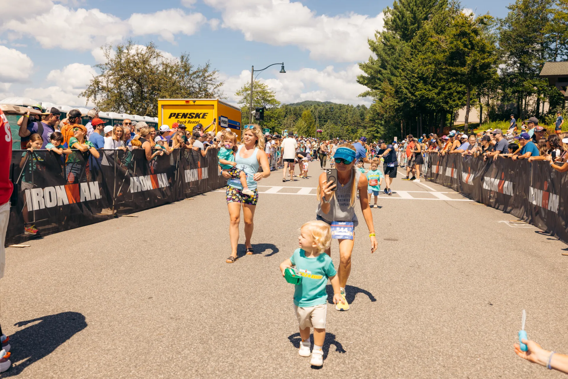 Woman walks behind child filming them and second woman carries toddler on Ironkids race course while spectators cheer