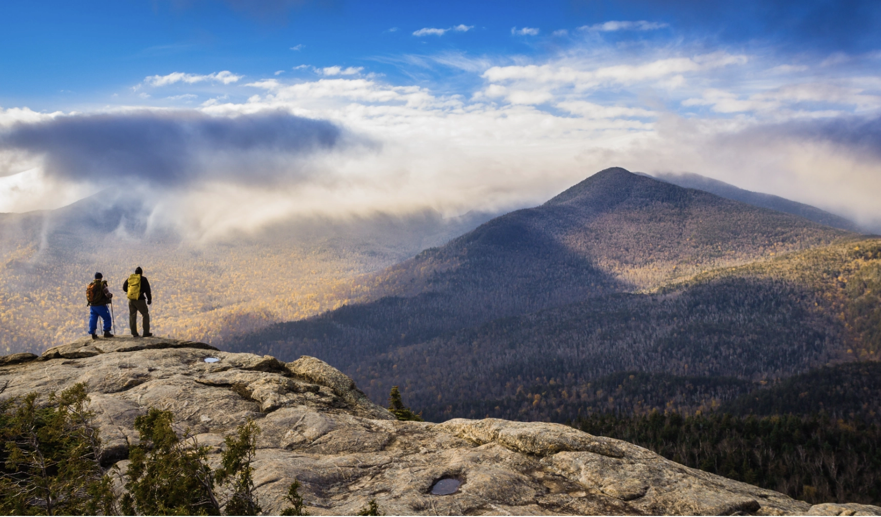 Two hikers stand atop a rocky mountain summit in the Adirondacks in fall looking out at view of other mountains