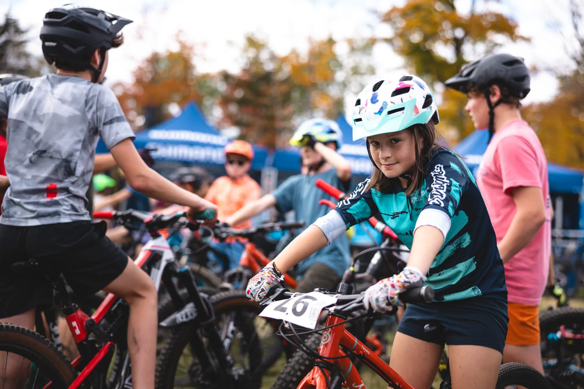 A young girl smiles while sitting on her mountain bike at a race, surrounded by other young racers.