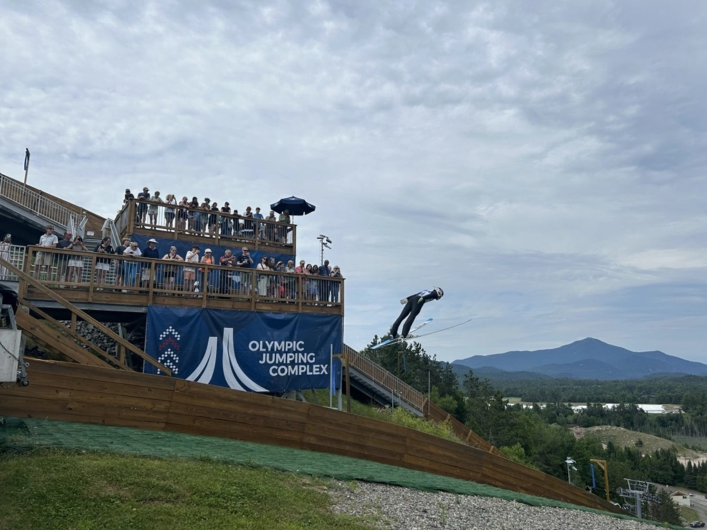 Ski jumper takes off from a lower jump over artificial turf while a two-level deck full of people looks on