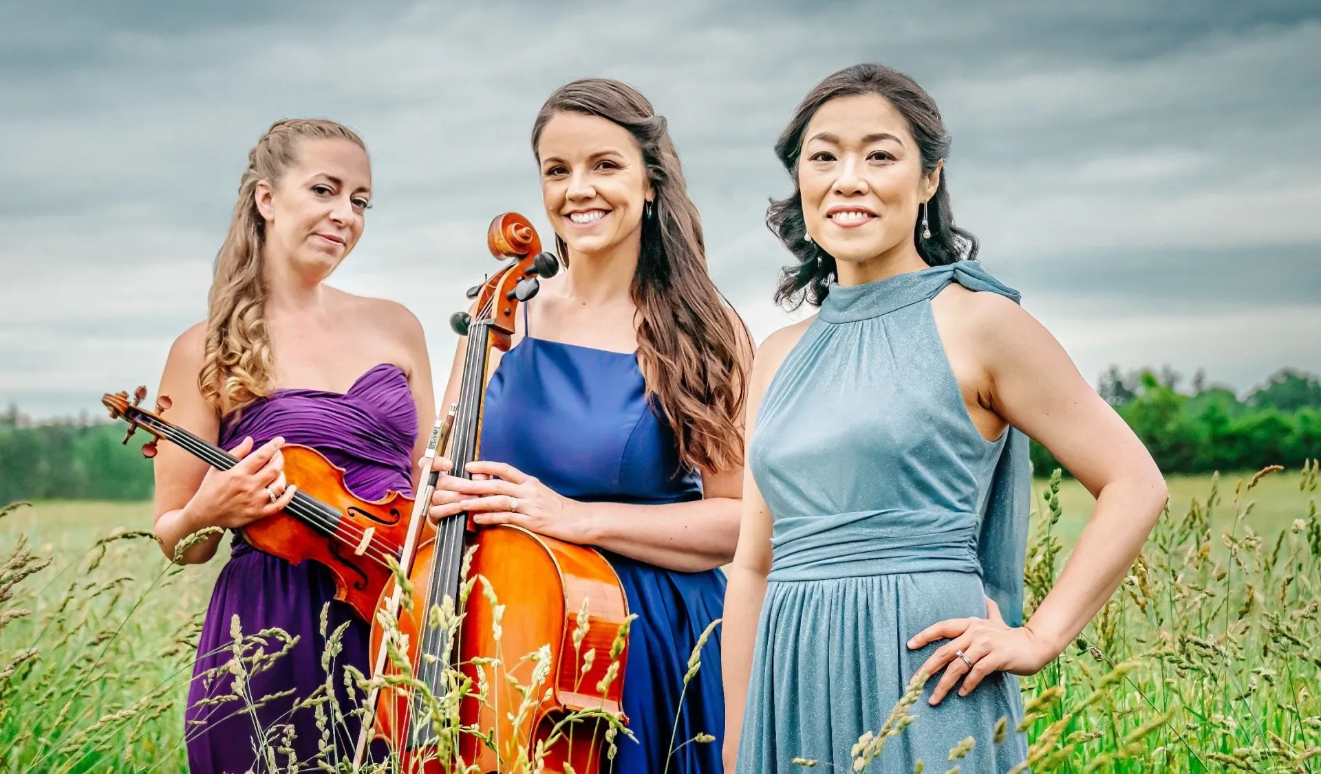 Three women in elegant dresses stand in a field. One holds a cello, another a violin.
