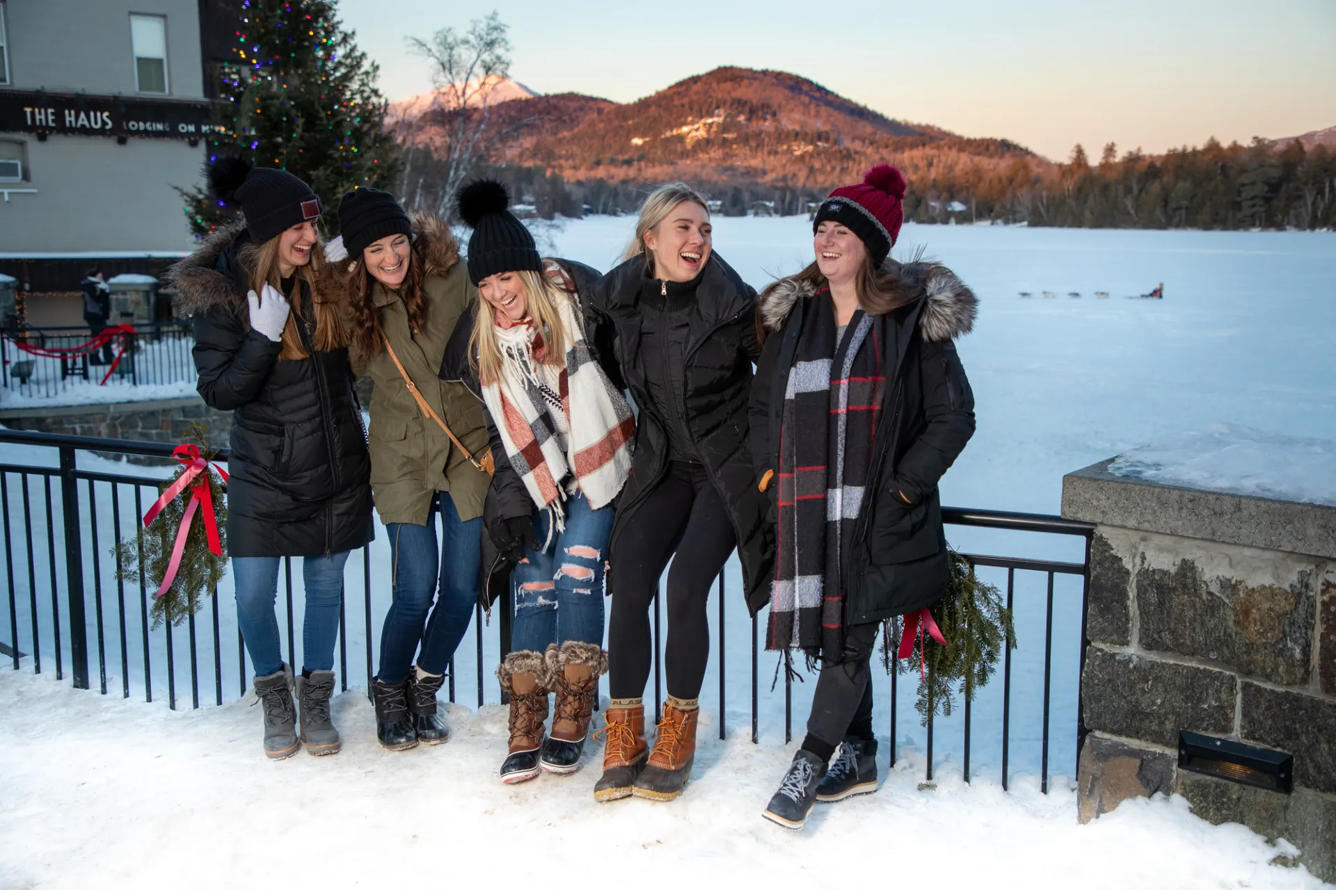Five women in winter clothing laugh in front of a snowy lake and sunset-lit mountains beyond.