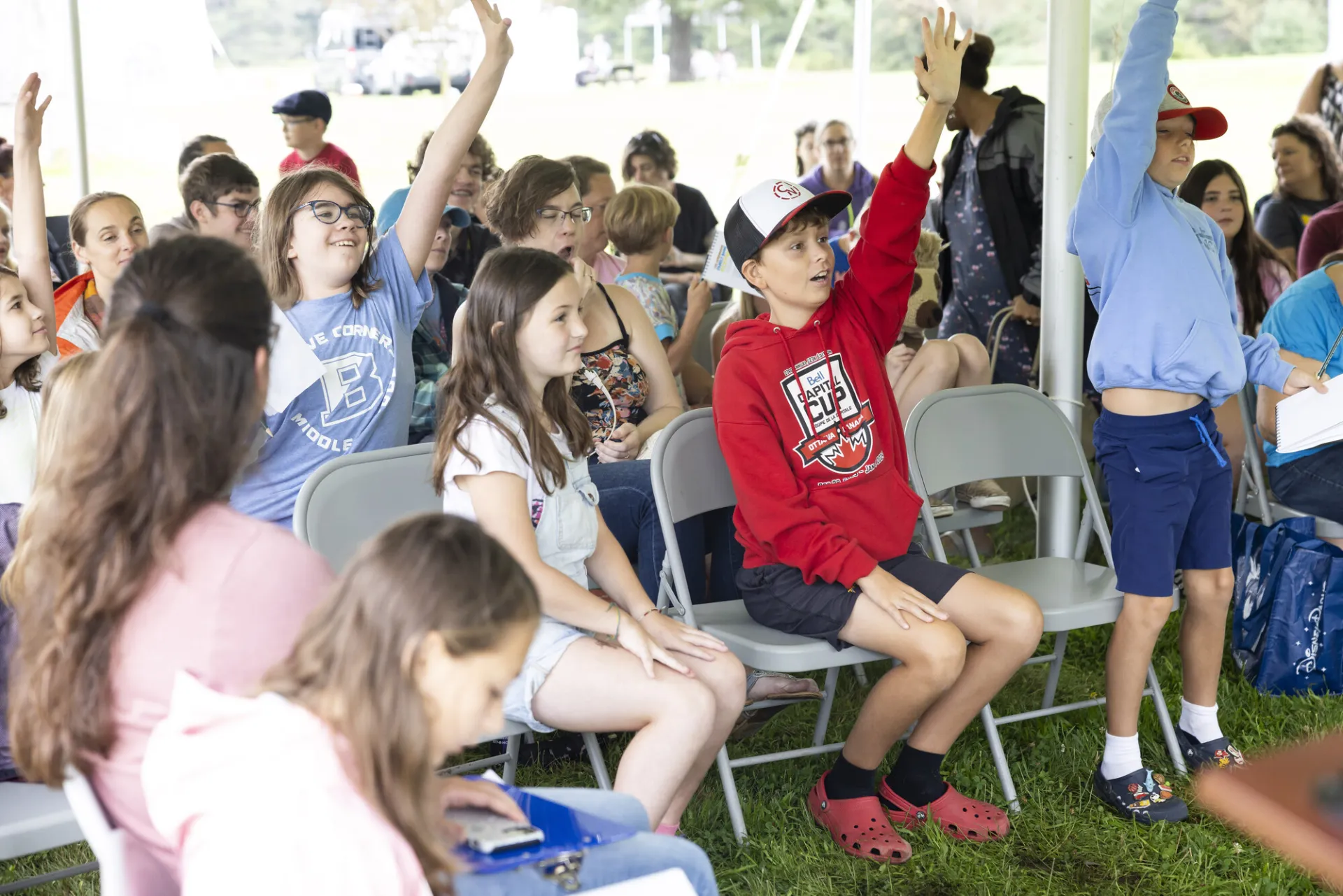 Collection of kids sit in folding chairs under outdoor tent at a presentation, some raising their hands