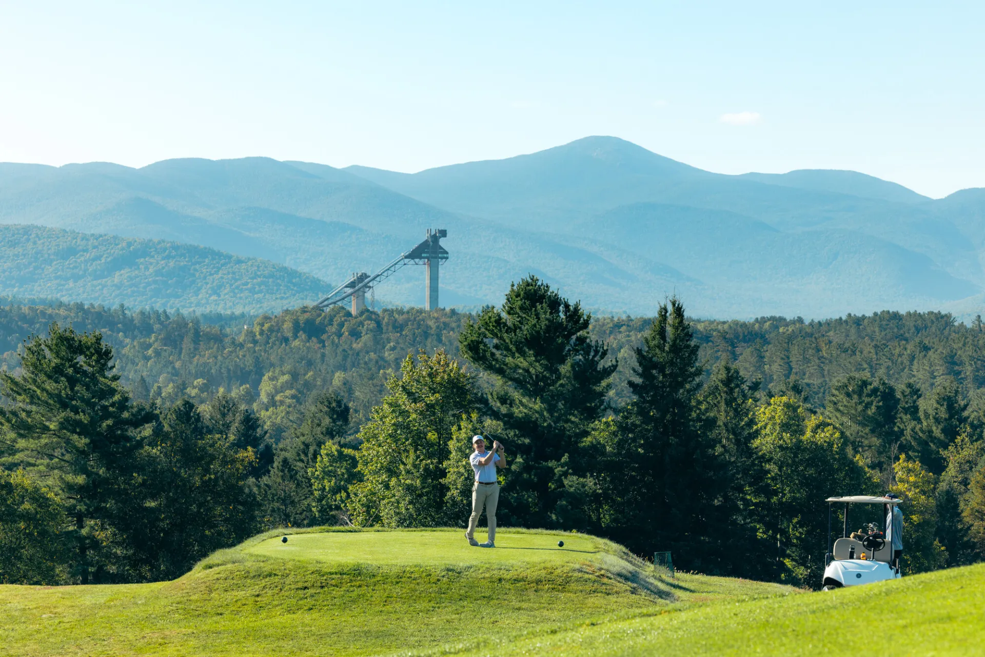 A man hits a golf ball in front of Olympic ski jumps. 