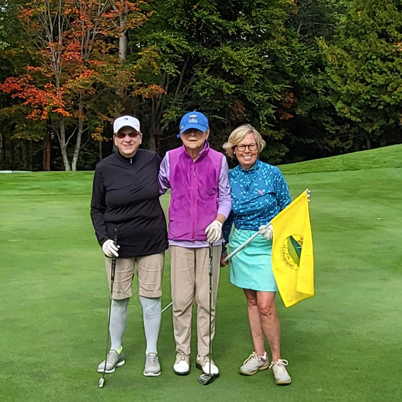 Trio of golfers stand together posing for camera on golf course, two holding golf clubs and one holding a yellow flag