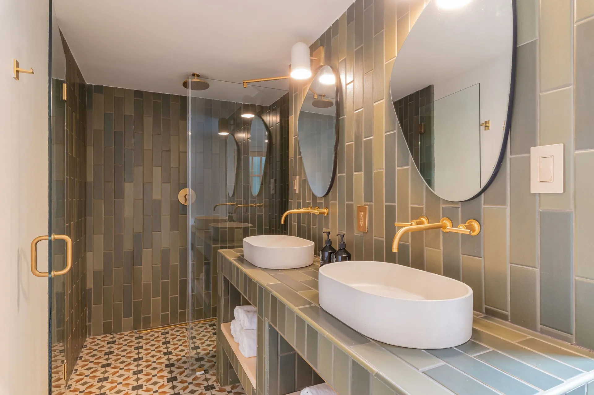 A bathroom with vertical subway tiles in earthy green hues, with walk in shower, his and hers basin sinks and organically shaped mirrors.