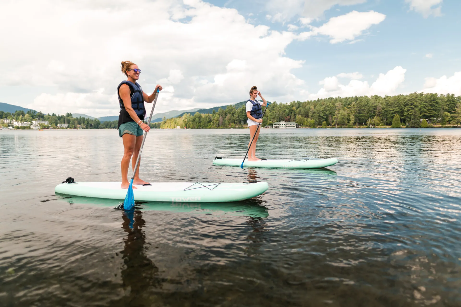 Two paddleboarders wearing personal floatation devices paddle on a gentle lake on a summer day.