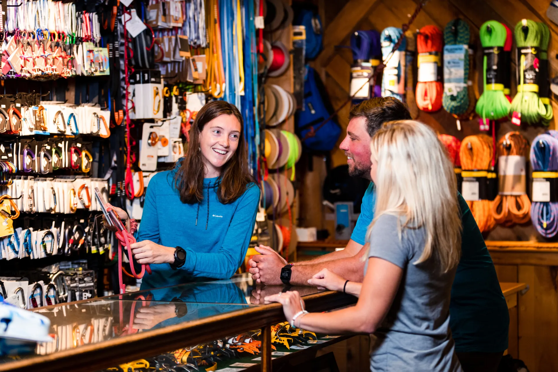 A smiling shop employee behind a glass counter shows a climbing rope to a man and a woman in the Mountaineer.