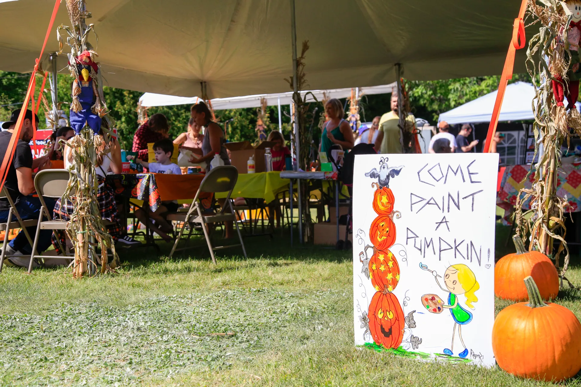 People sit under an outdoor tent with children with a sign out front that says "come paint a pumpkin"