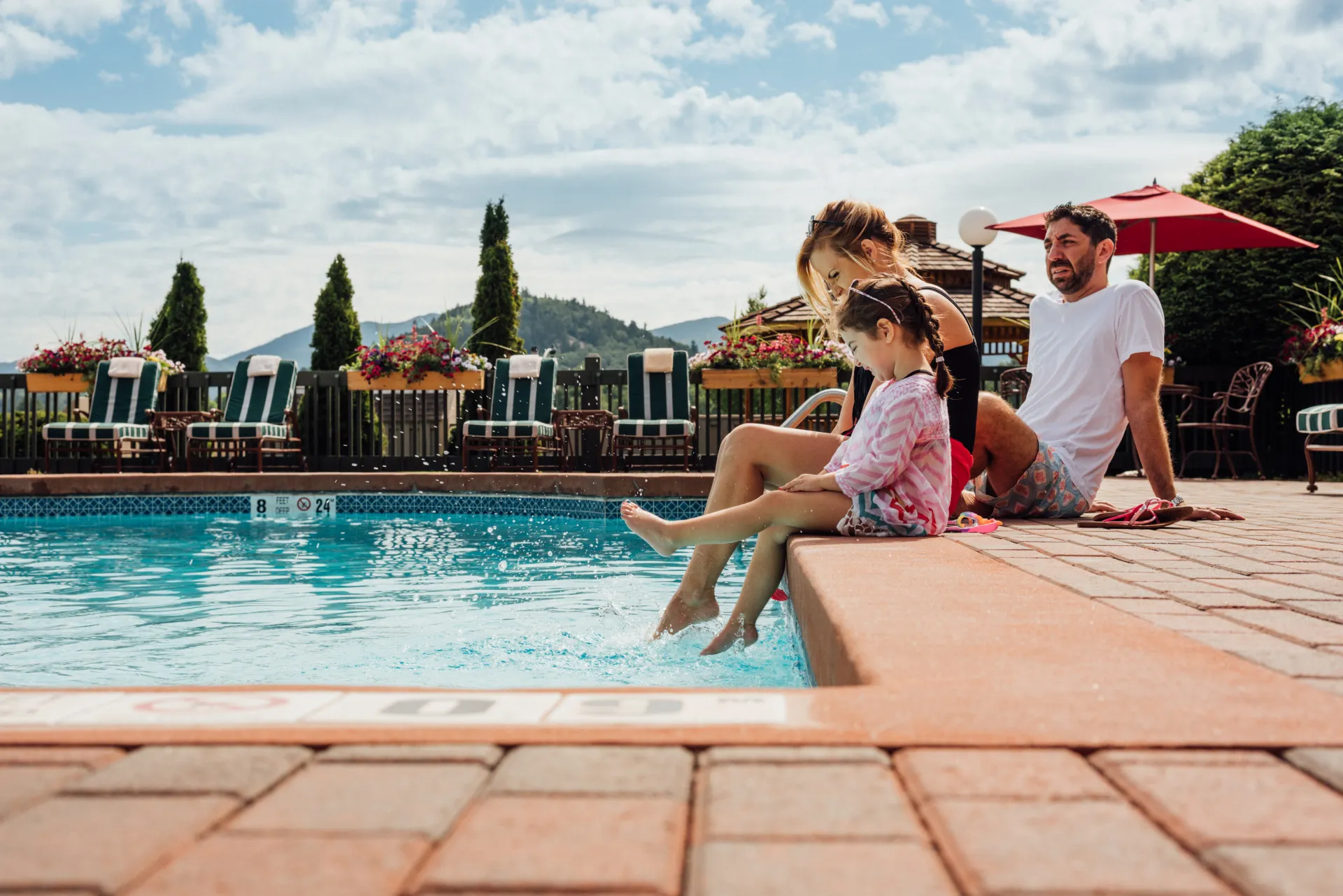 Small girl and parents sit on edge of pool putting their feet in the water in summer