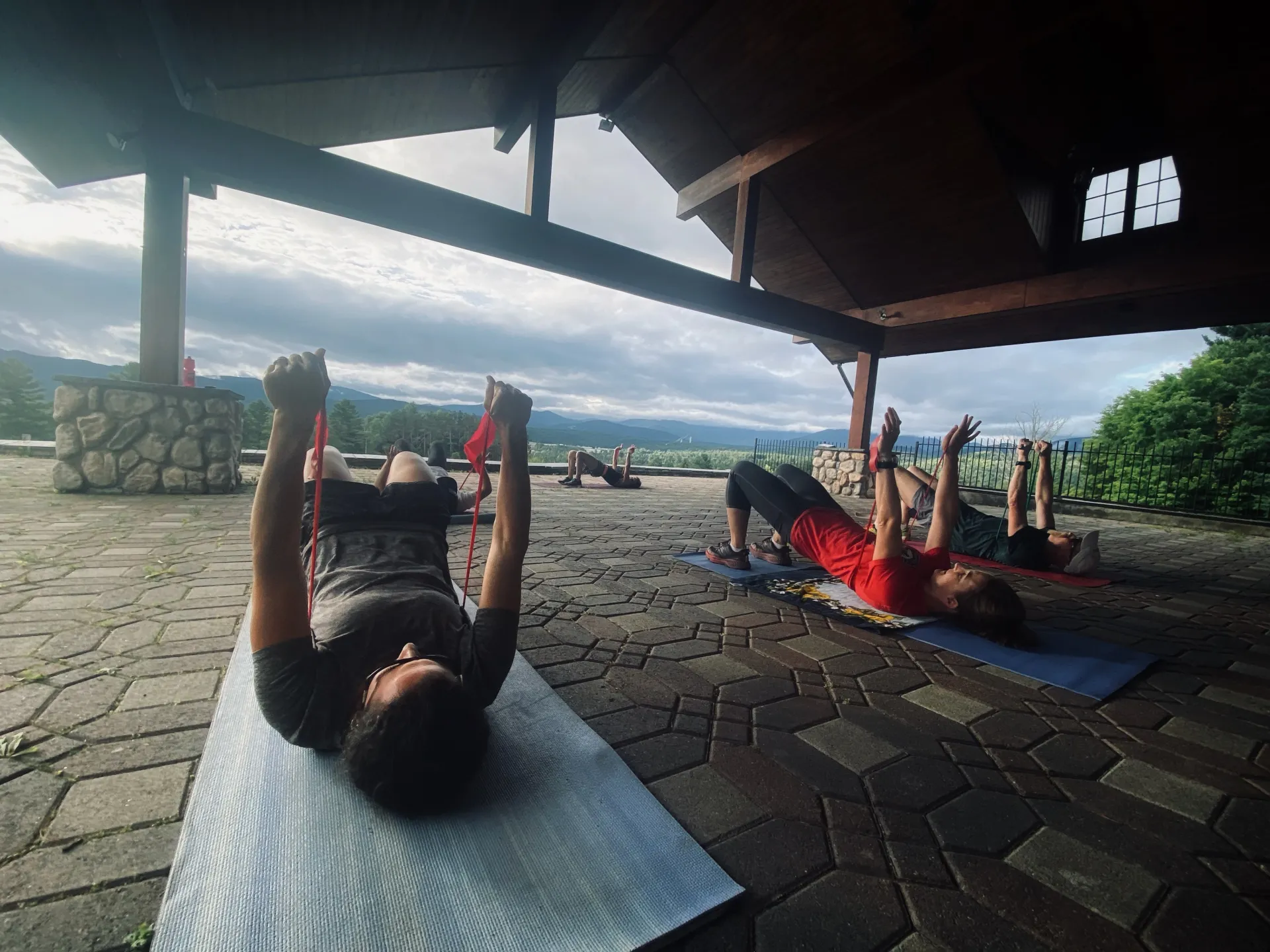 People lay on backs on yoga mats pushing arms up with resistance bands in workout class held outdoors on covered brick patio