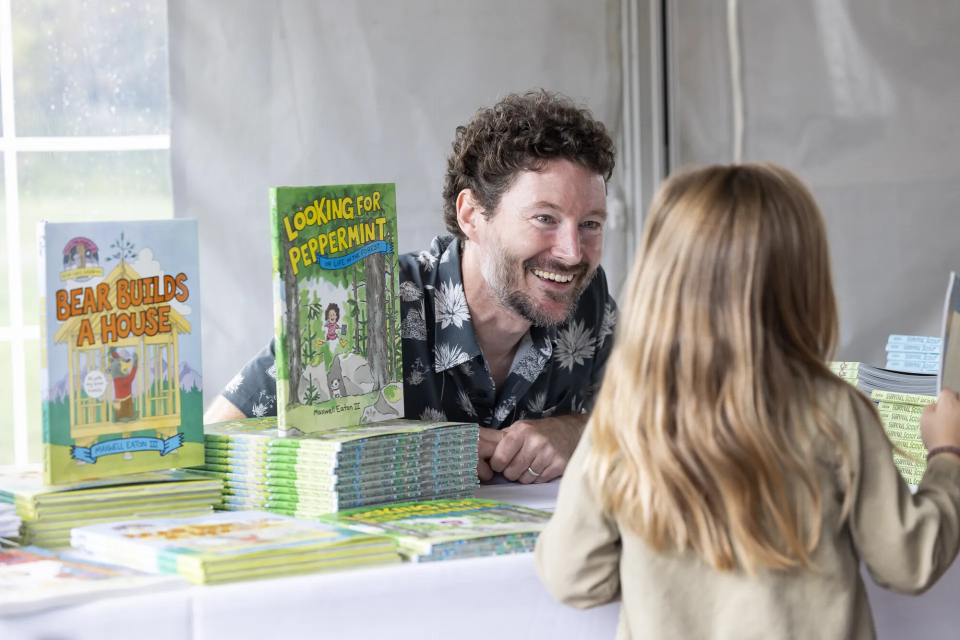 Middle aged white male author sits behind book display on table smiling at child standing across from him