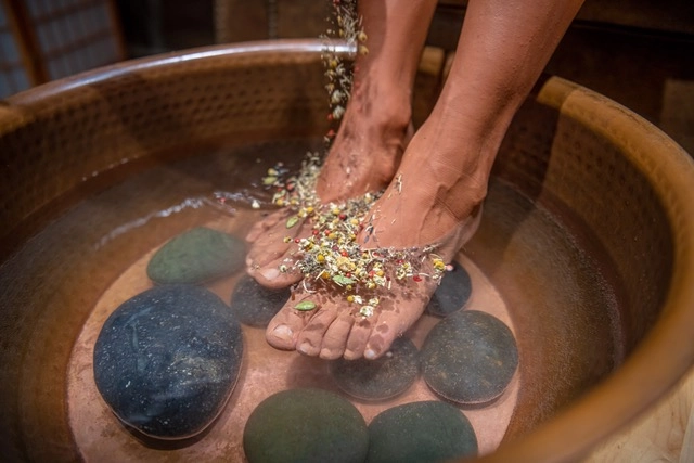 Feet immersed in a tub of steaming water, topped with herbs and some river stones within