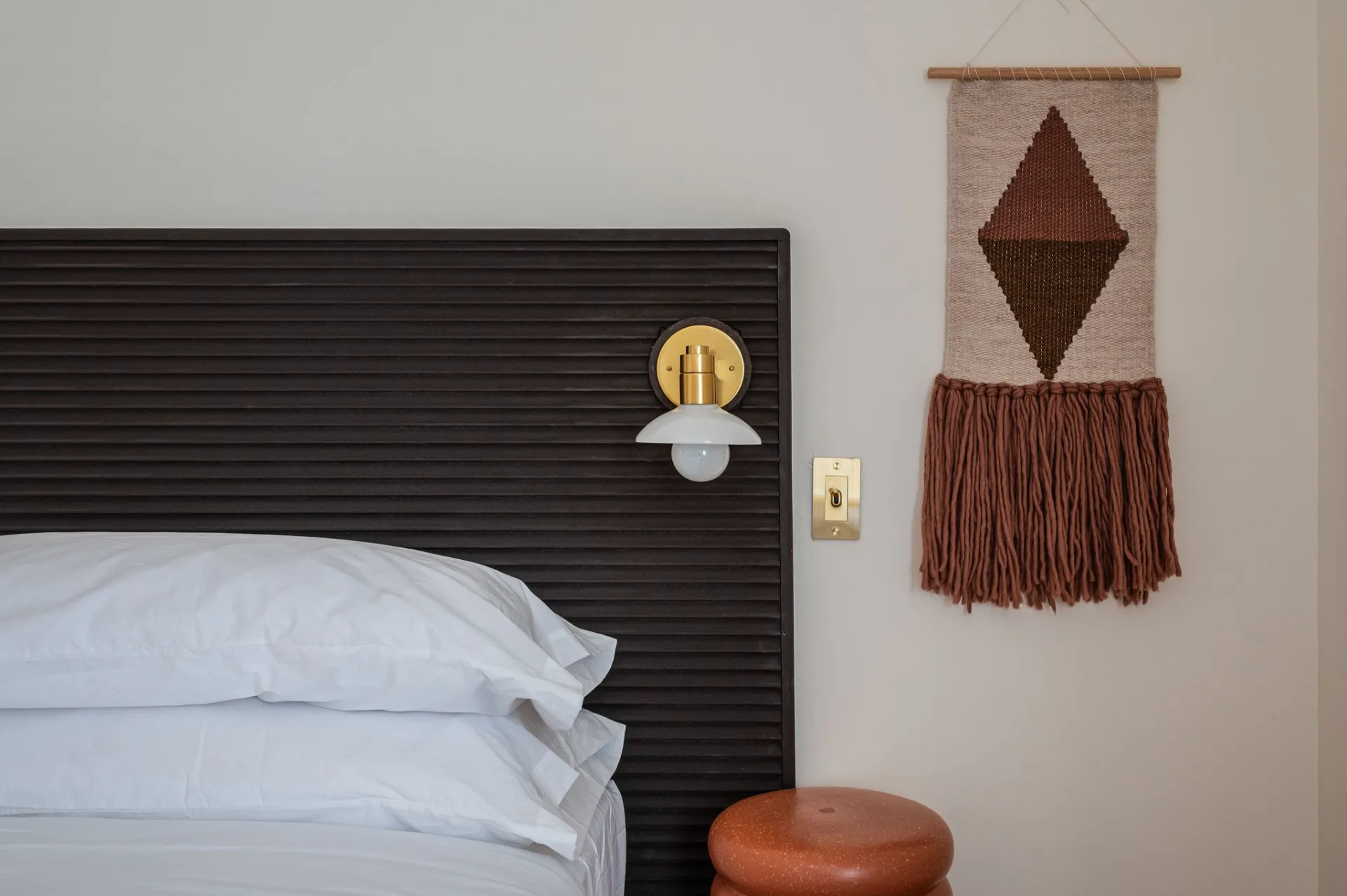 A rust and oatmeal colored woven wall hanging next to the brass wall scone and minimalist night stand at Eastwind Lake Placid.