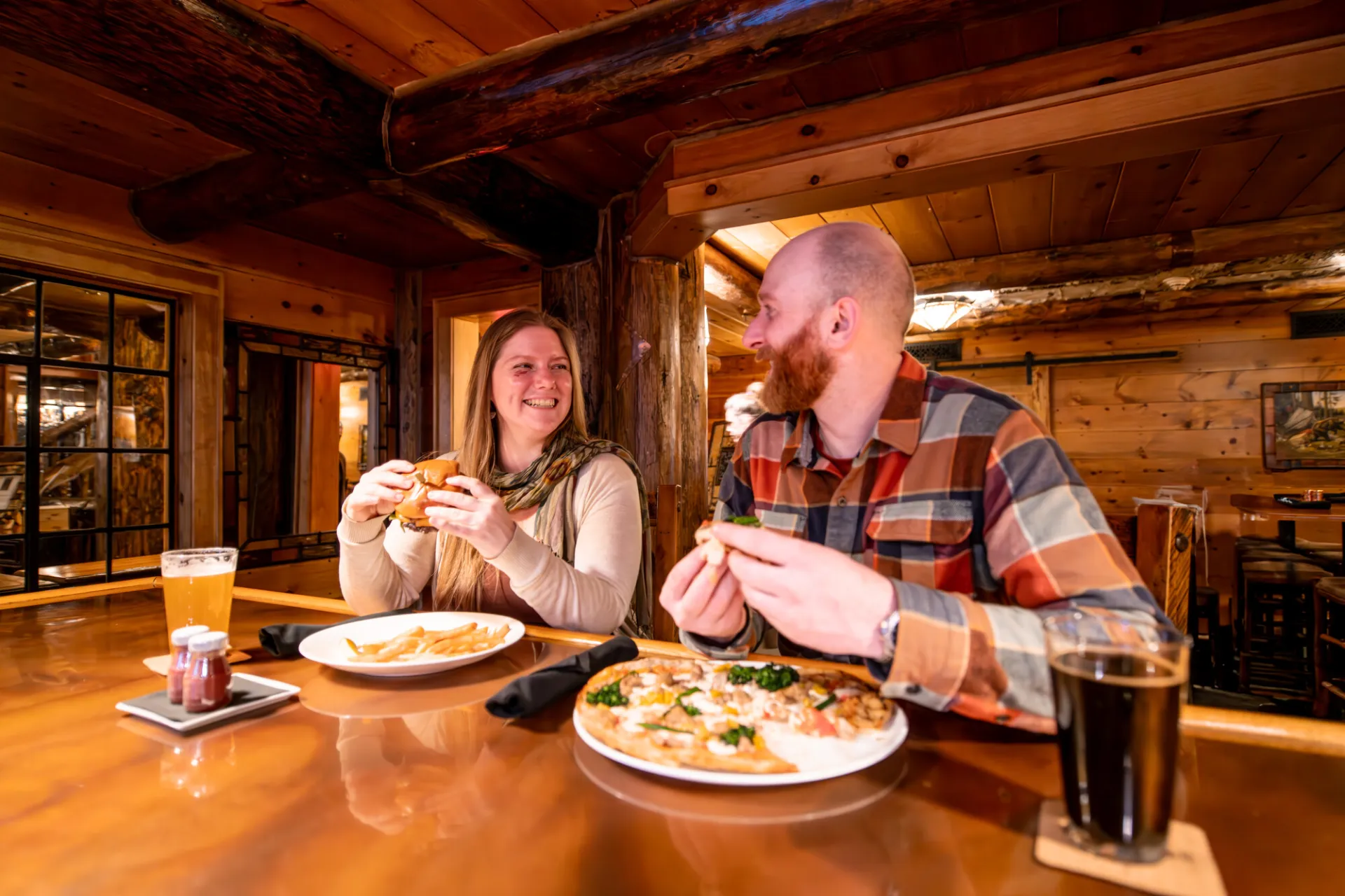 Man and woman sit side-by-side at bar smiling at one another with pints of beer and plates of food holding burgers