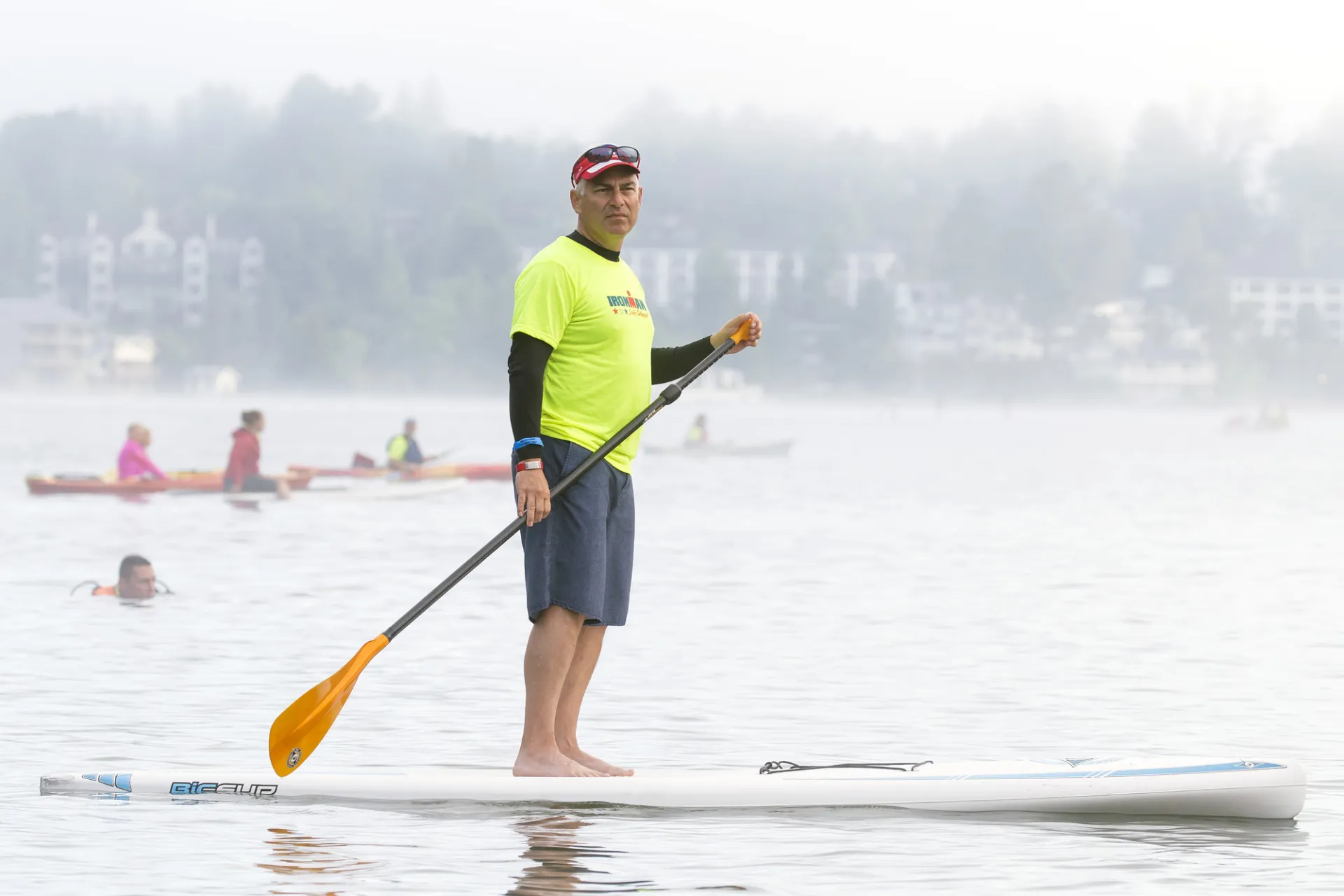 A man in a bright yellow Volunteer tshirt and shorts stands on a paddle board on a misty morning watching a race